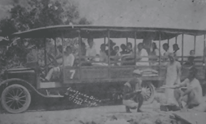 A circa-1920 public transportation vehicle carrying passengers, with several people gathered beside it.