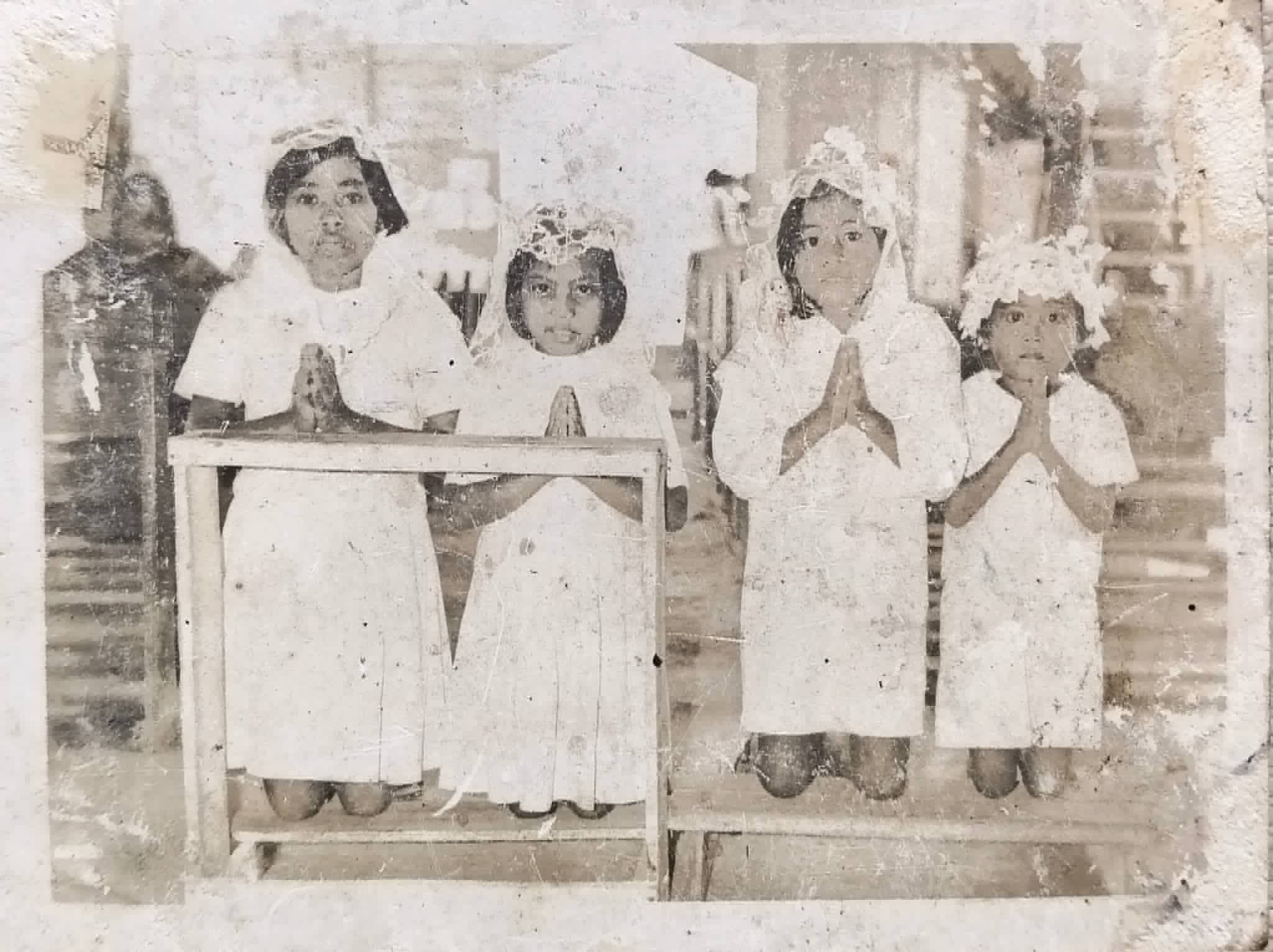 Four children in white ceremonial clothing kneel with their hands joined in prayer.