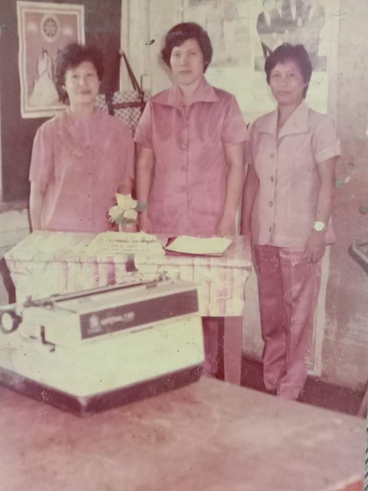 Three women stand behind a desk with a typewriter inside a wooden office.