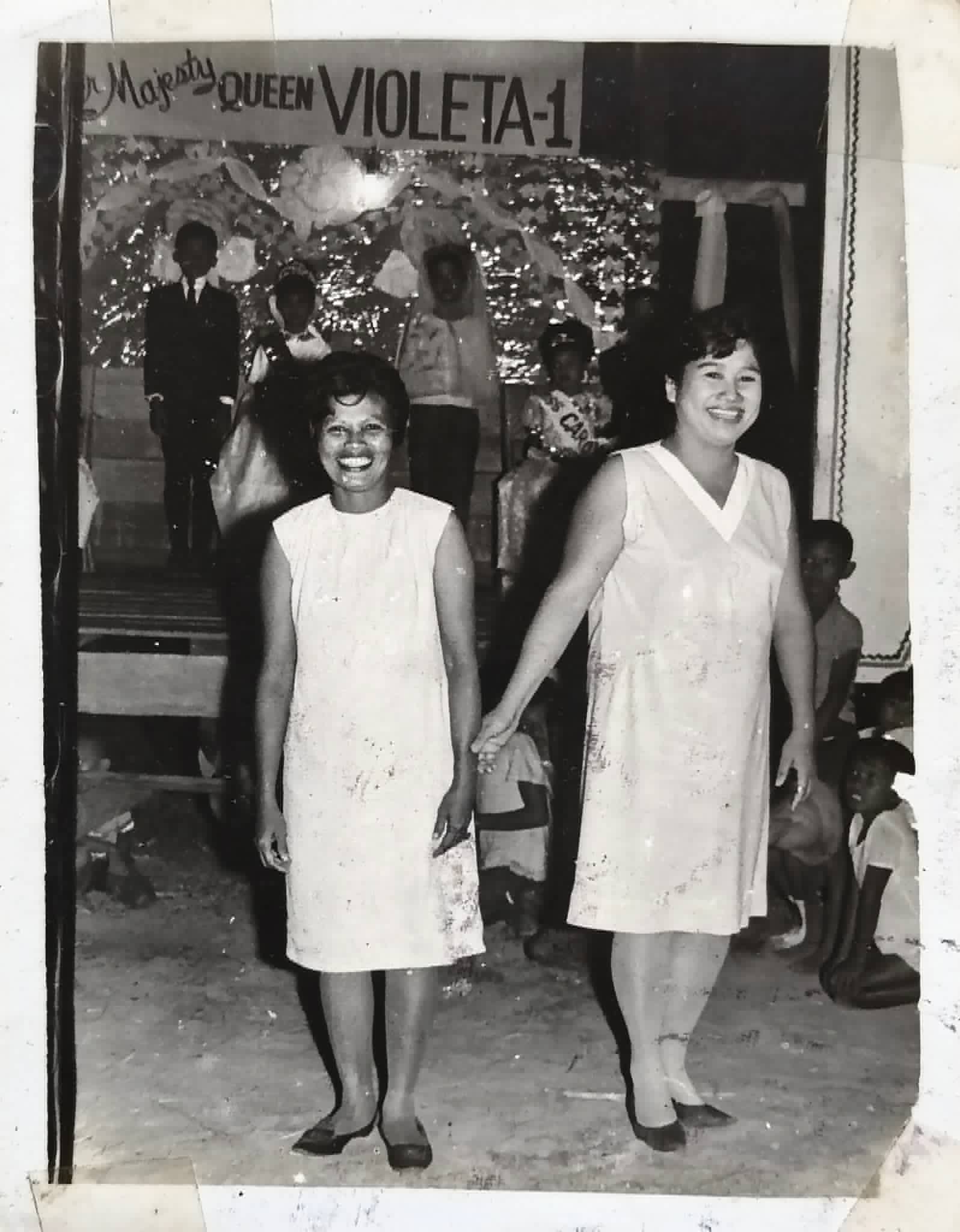 Two women walk hand in hand in front of a decorated stage marked Queen Violeta-1.