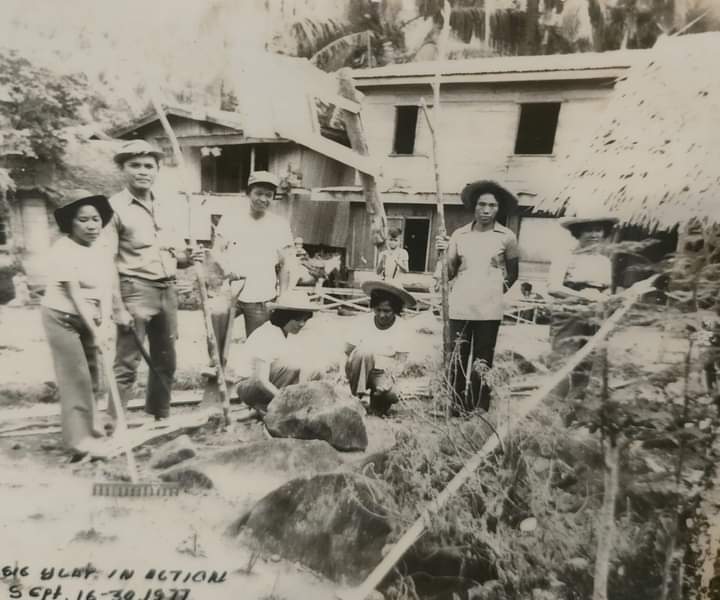 Six people stand and crouch among rocks and construction materials at a work site in 1977.
