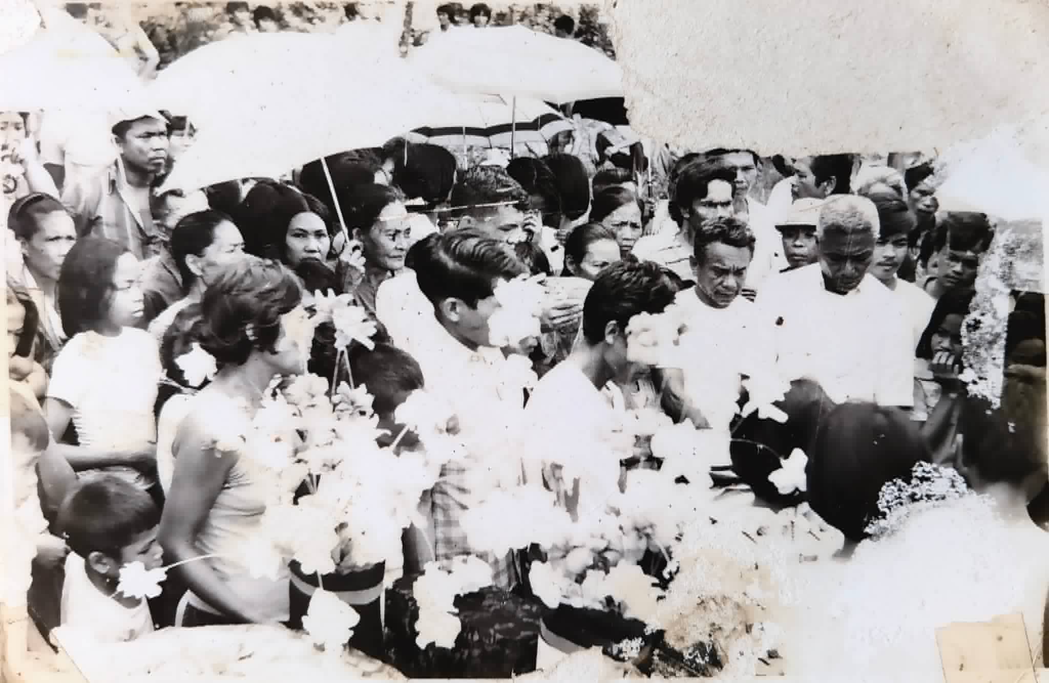 A large crowd gathers around several people holding flower bouquets under umbrellas.