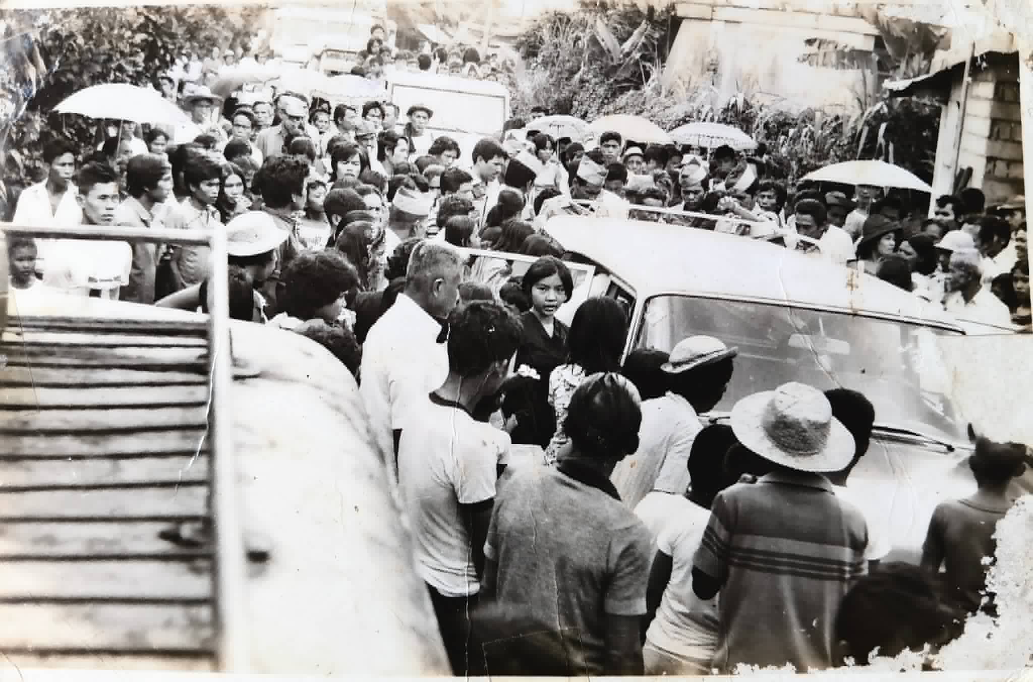A large crowd surrounds a motor vehicle on a busy street.