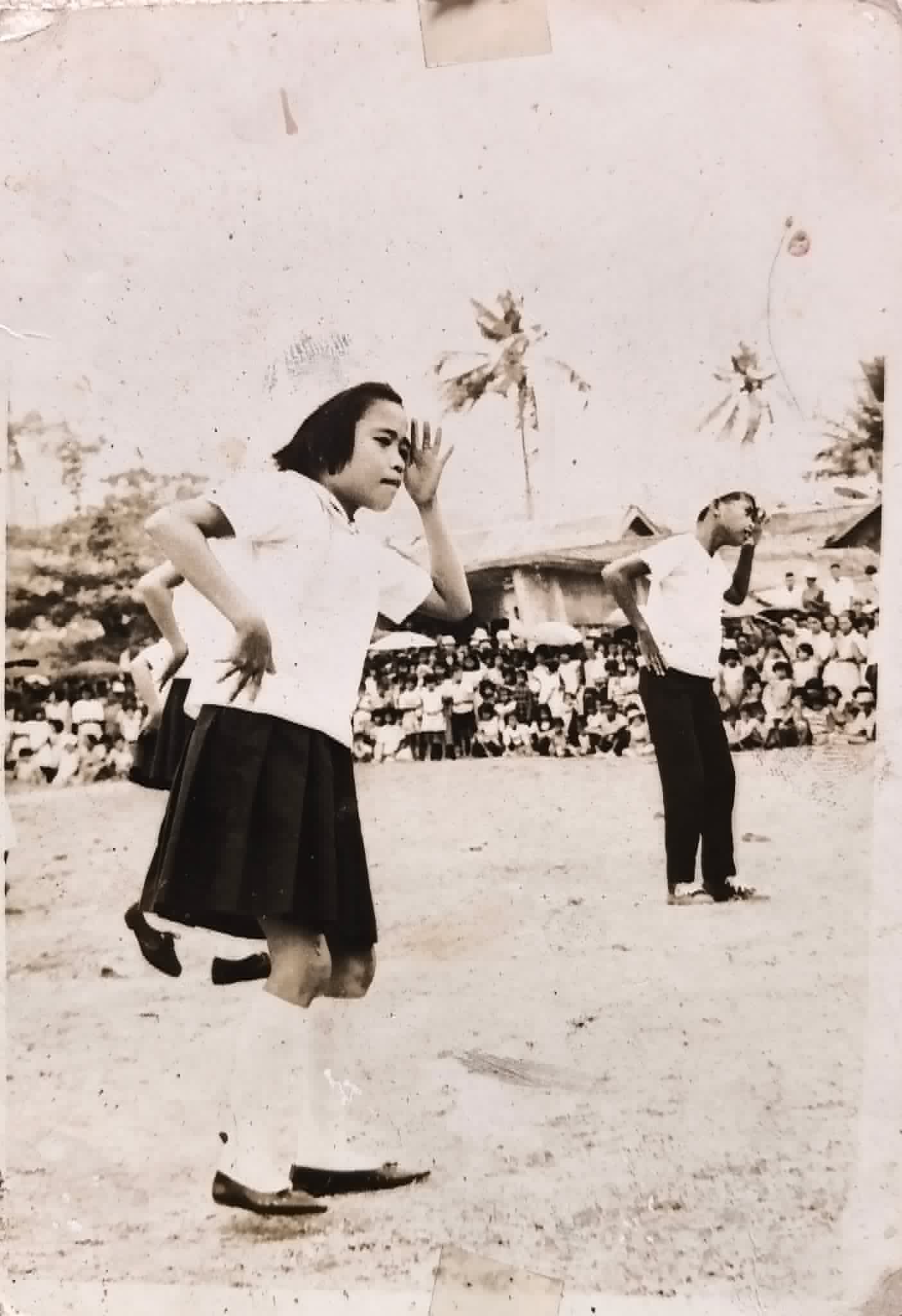 Young performers dance in an open area while a large audience watches.