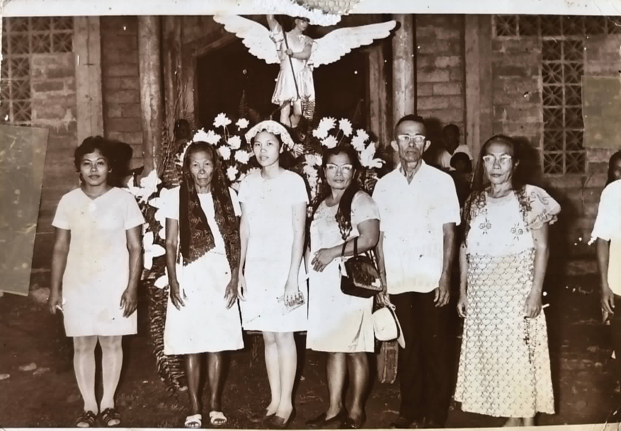 Six adults pose in front of a flower-decorated religious image of a winged figure.
