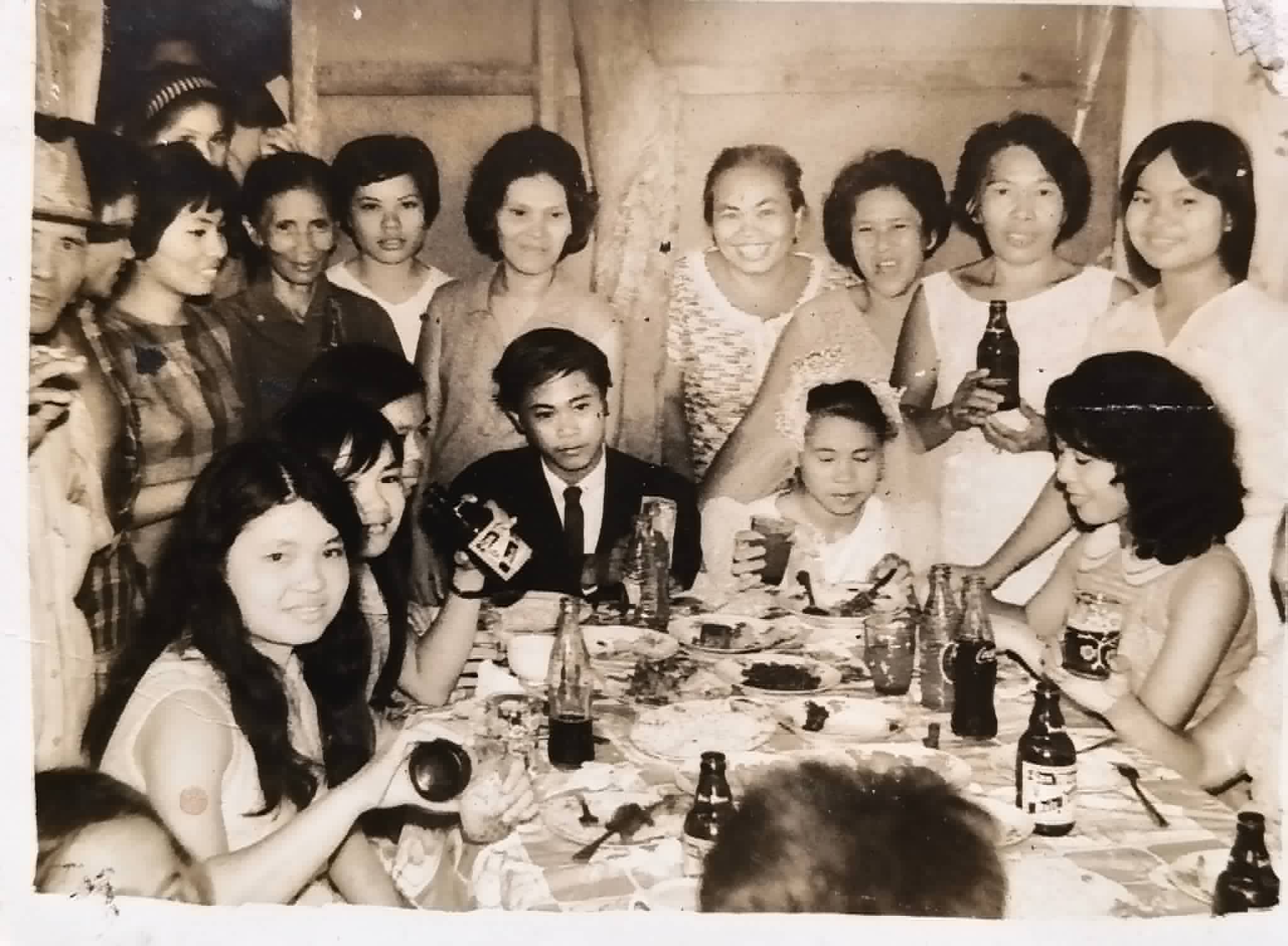A large group of adults and children gathers around a table with food and bottled drinks.
